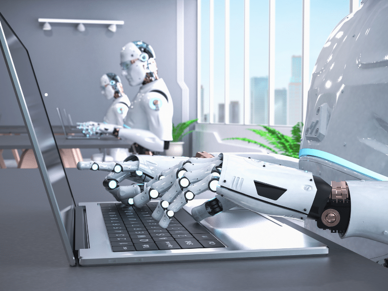 AI robots sat at a grey desk typing on a keyboard of a laptop