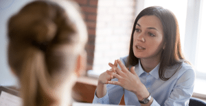a woman with long brown hair and wearing a blue button up shirt is having a discussion with a team member. her hands are interlocked